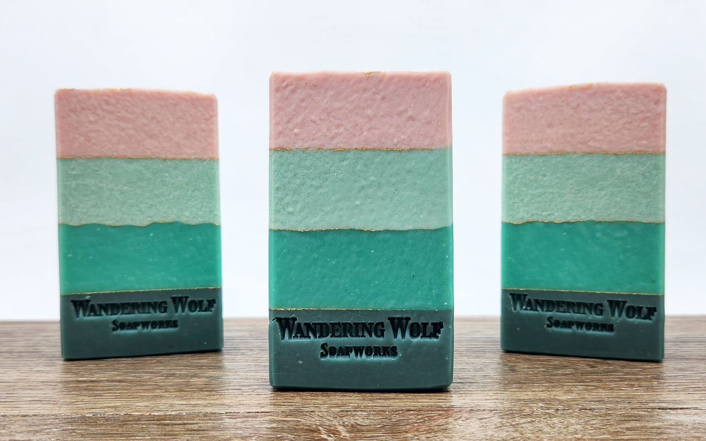 Three bars of soap with layered colors of pink, green, and teal, each marked with the brand 'Wandering Wolf Soapworks'.